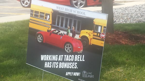 Taco Bell sign - "Working at Taco Bell has its bonuses"; includes image of teenage boy standing in front of school buses showing a teenage girl his red convertible